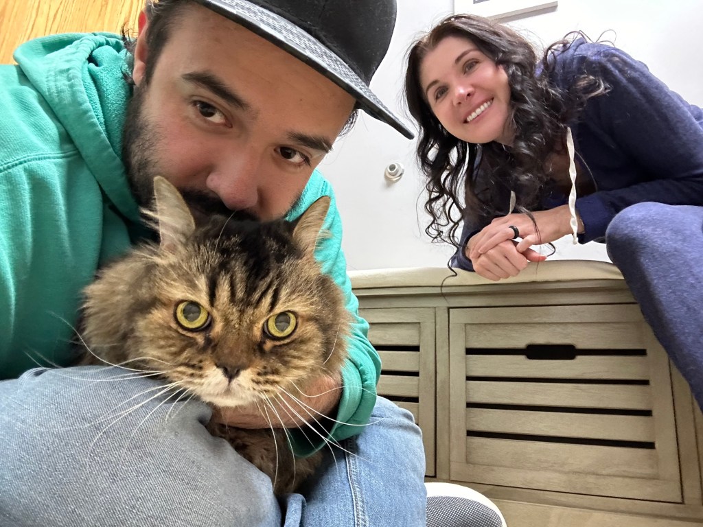 Zach, owner, poses while holding Max, a gray tabby, and Maria, owner, in the background also posing. 